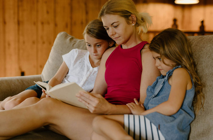 A woman in a red top reads a book on a couch with two young girls, showcasing family time.