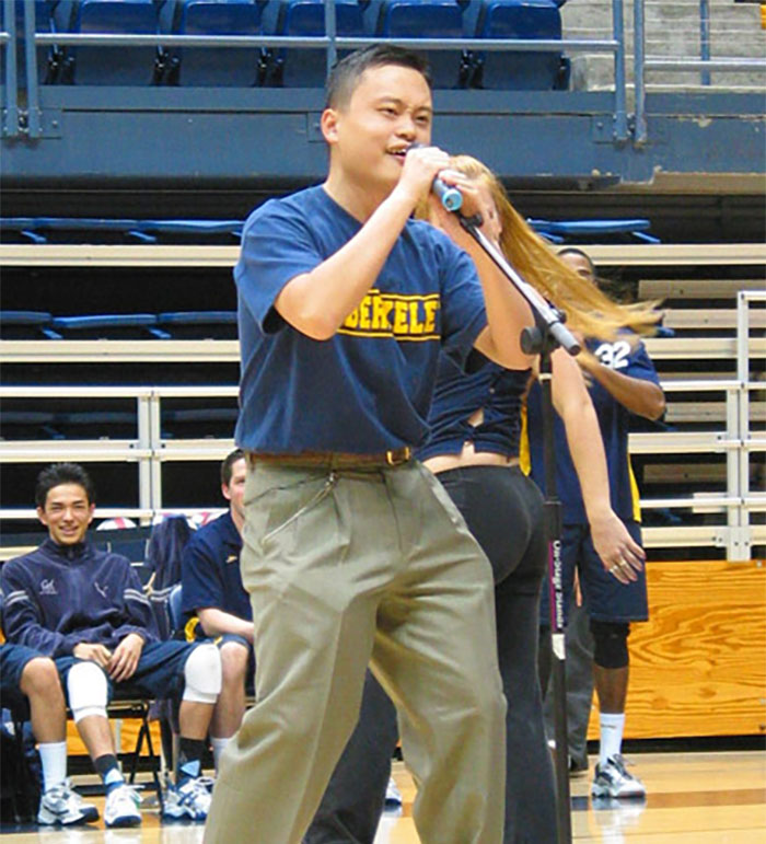 Man singing passionately into a microphone on a basketball court, wearing a Berkeley shirt.