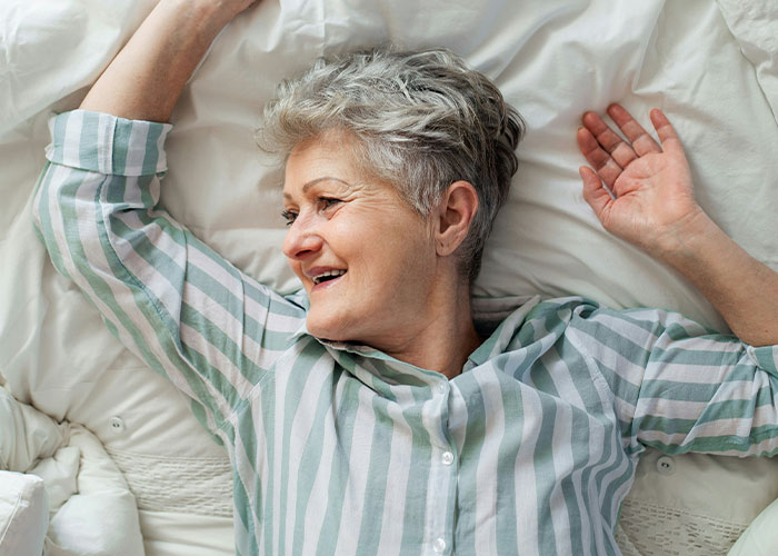 Elderly woman in striped pajamas smiles lying in bed, conveying heartfelt emotions.