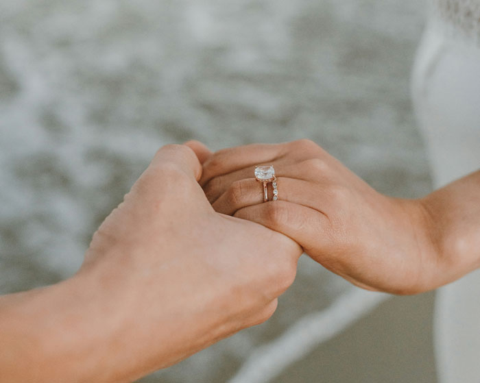 Two hands holding each other, one wearing an engagement ring, with a beach background; uncovering a man's background theme.