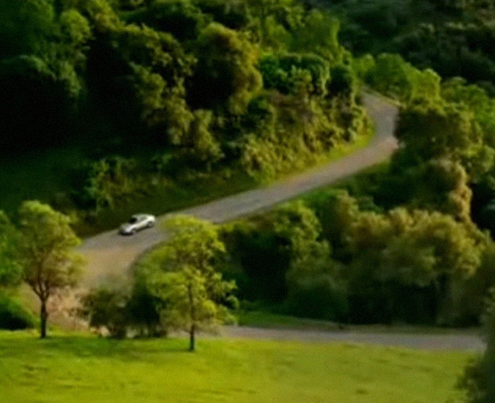A 2000s car drives on a winding road surrounded by lush greenery, evoking nostalgic memories.