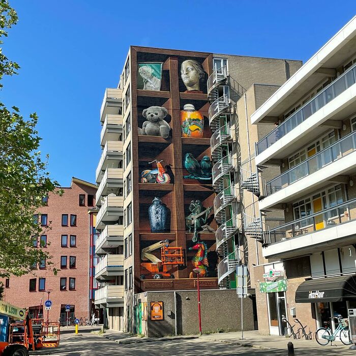 Mural by Jan Is De Man depicting hyperrealistic bookshelves on a city building wall.