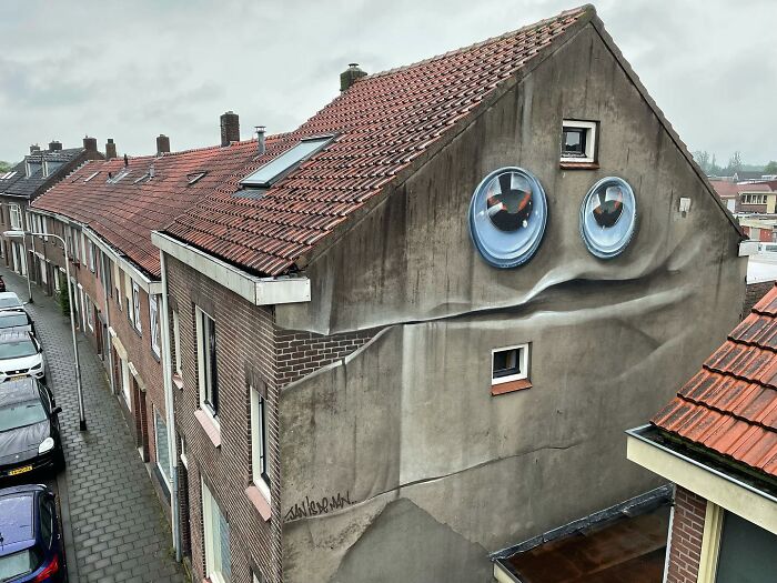Hyperrealistic mural by Jan Is De Man on a city building, depicting a face illusion with windows as eyes.