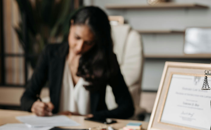 Woman in an office setting writing on papers, with a framed certificate visible, representing the theme of prettiest girl from HS.