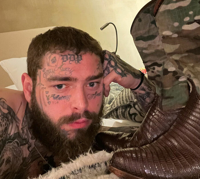 Bearded man with tattoos lying on a bed beside camo-print clothing, contemplating something about a celebrity.