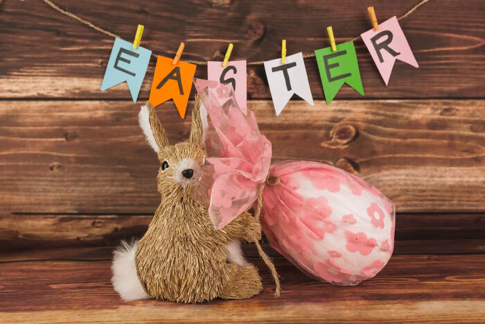 Easter bunny decoration holding a floral-patterned wrapped egg under colorful Easter banner on wooden background