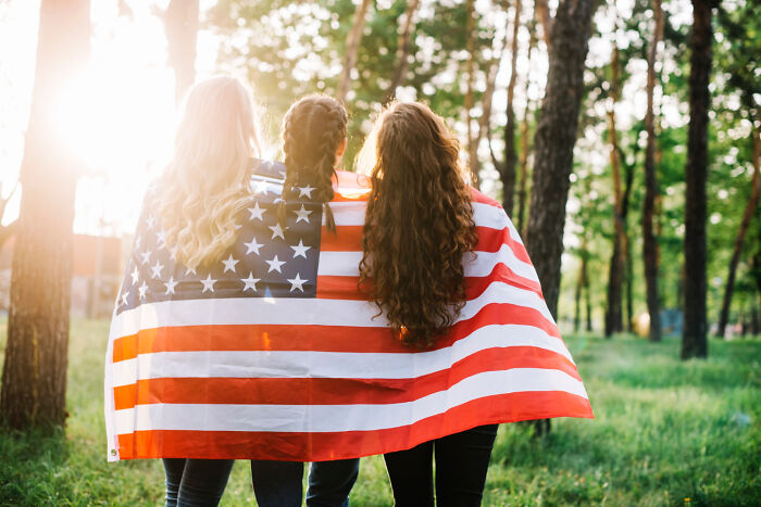 Three women wrapped in an American flag, standing in a sunlit forest, symbolizing American norms.