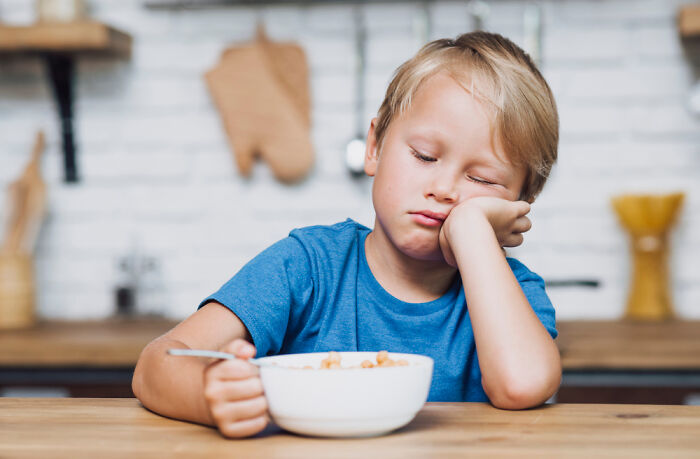 Young boy in a blue shirt looking tired and uninterested while sitting at a table with a bowl of cereal, illustrating uncommon dessert experiences.