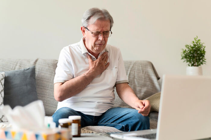 Elderly man sitting on couch holding chest, appearing unwell, surrounded by medication bottles and tissue box indoors.