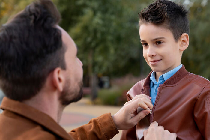 A father helping his son zip up a jacket outdoors, representing experiences people were raised to think were common.
