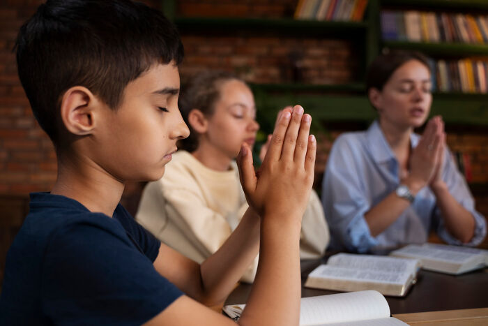 Three children with eyes closed and hands clasped in prayer, reflecting diverse experiences people were raised to think were common.