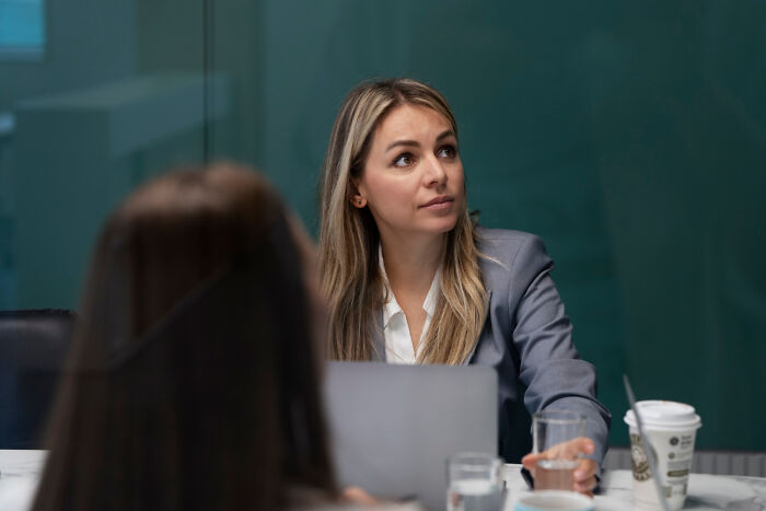 Woman in a gray blazer at a meeting, looking thoughtful, with a coffee cup on the table.