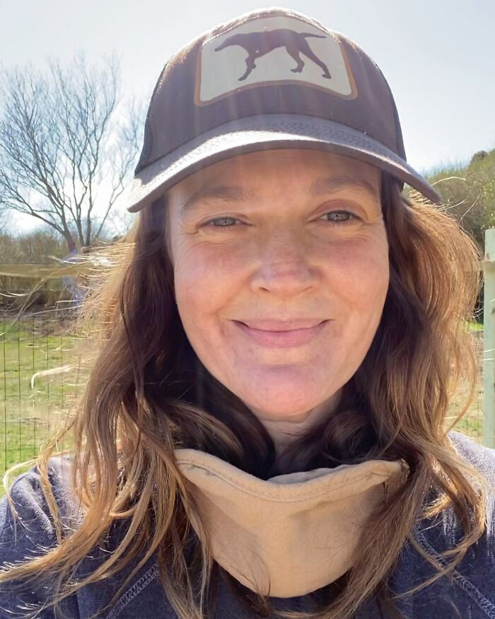 Celebrity posing without makeup, wearing a hat with a dog design, smiling outdoors in natural light.