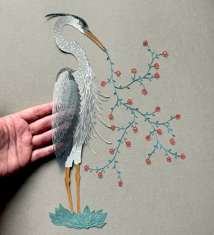 Intricate paper art of a heron by Pippa Dyrlaga, featuring detailed feathers and floral elements, held in a hand.
