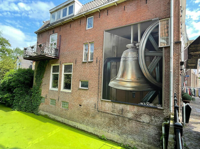 Hyperrealistic mural by Jan Is De Man on a city wall, depicting a detailed bell with intricate shadows and reflections.
