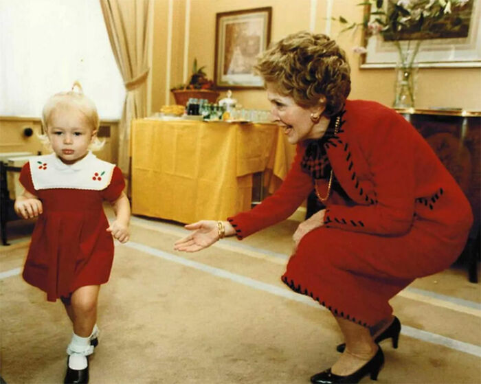 Woman in red suit kneeling, smiling at a toddler in a red dress, illustrating a rare historical moment indoors.