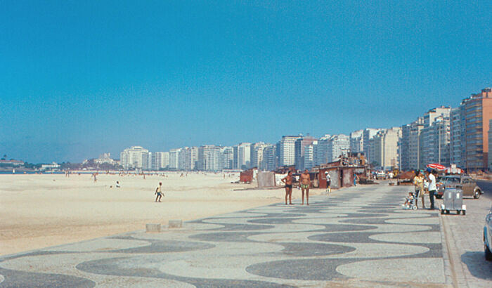 Retro Brazilian beach scene with sun and sand, featuring a curved promenade and distant high-rise buildings.