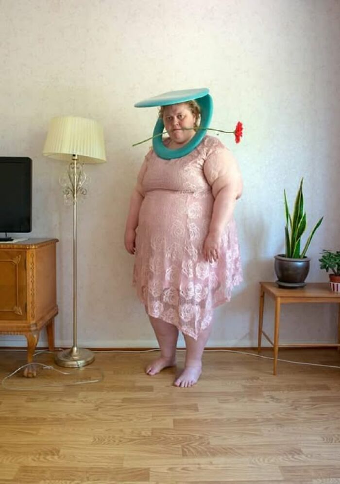 Person in a pink dress with a toilet seat around their neck, holding a flower in mouth; a weird image in a casual setting.
