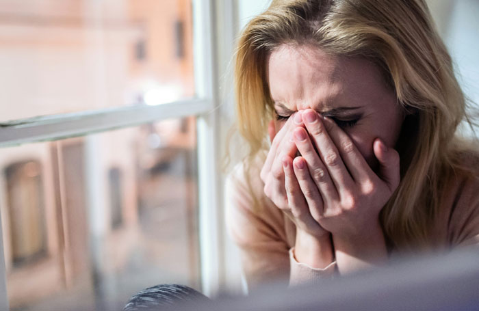 A blonde woman sitting by a window, covering her face with her hands, looking emotional.