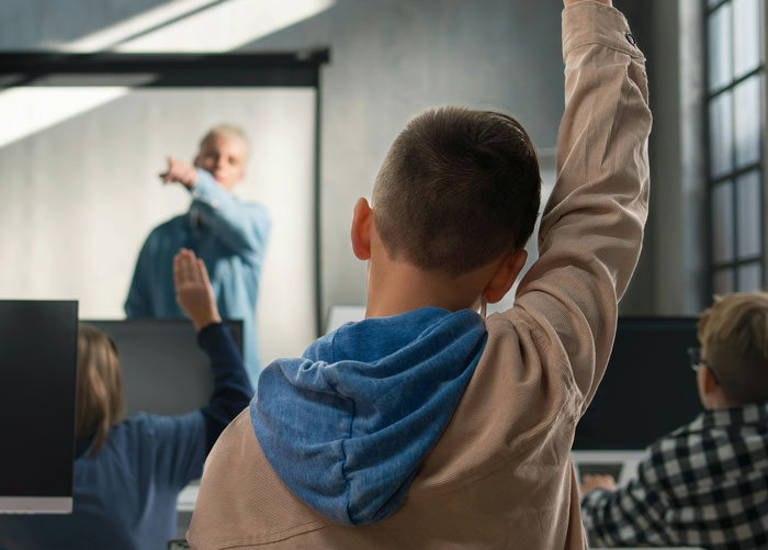 Estudiantes en clase, uno levantando la mano mientras el profesor parece sonreír.