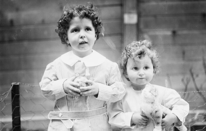 Two children with curly hair hold toys, dressed in vintage clothing, representing rare historical photos.