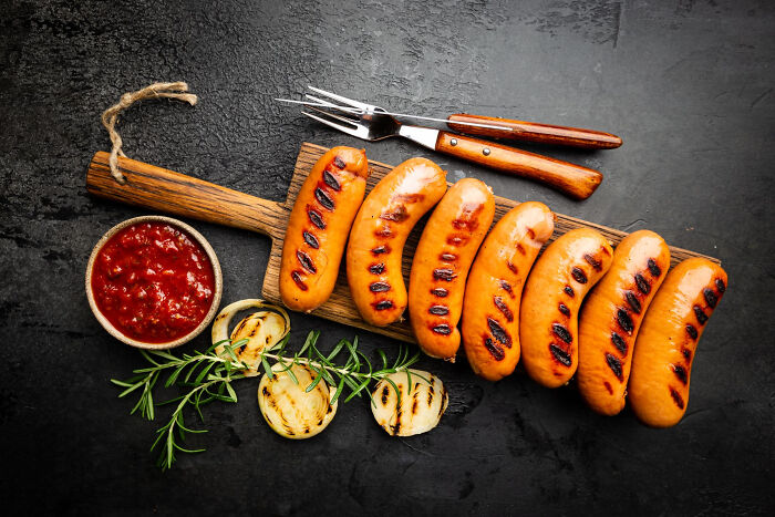Grilled sausages served on a wooden board with roasted onions, rosemary, and a bowl of red sauce, illustrating common food experiences.