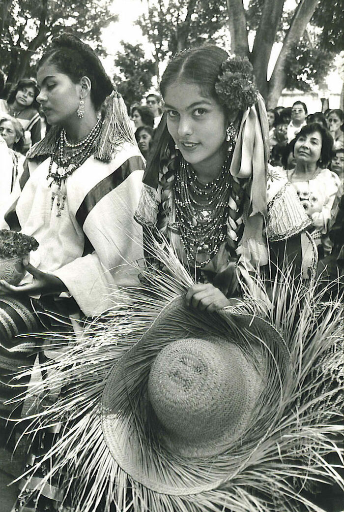 Two women in traditional attire holding woven hats, surrounded by a crowd, captured in a rare historical photo.