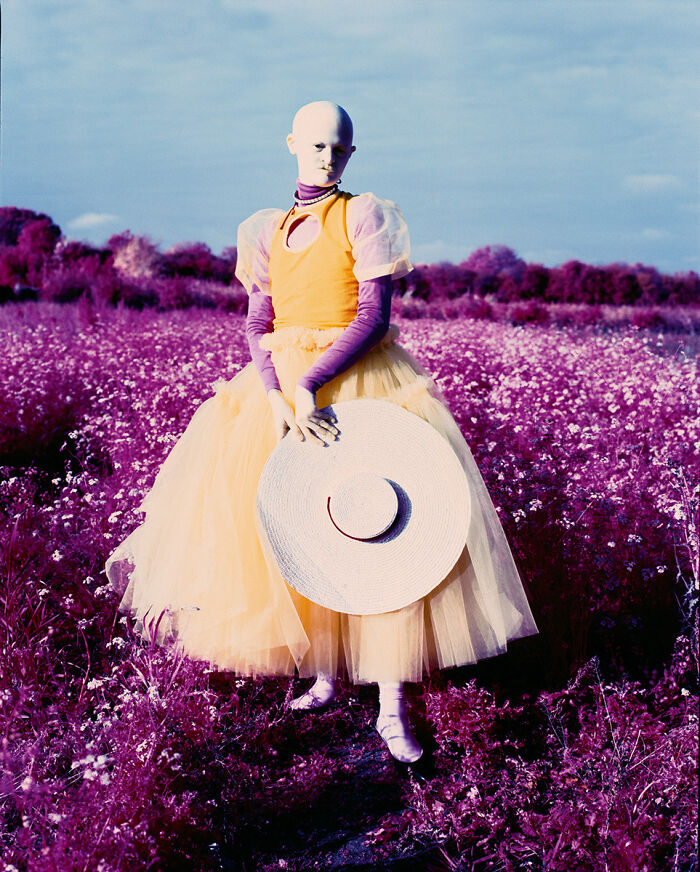 Unique model in colorful field, wearing layered dress and holding large straw hat, showcasing captivating style.