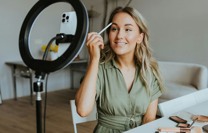 Young woman in a green dress applying makeup in front of a smartphone and ring light, symbolizing beauty and self-expression.