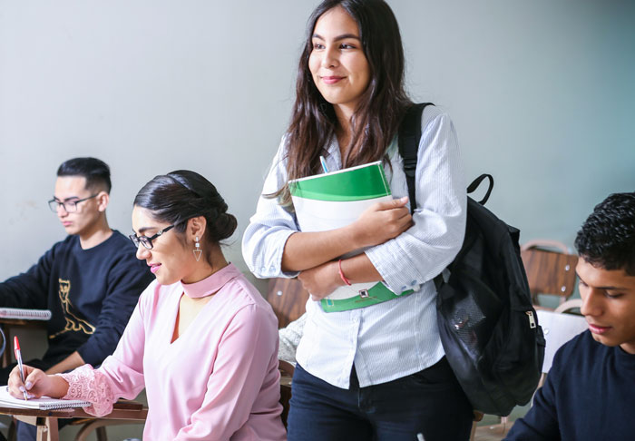 Estudiantes en clase, uno saliendo al frente con una mochila y cuadernos, provocando risas entre compañeros y profesores.
