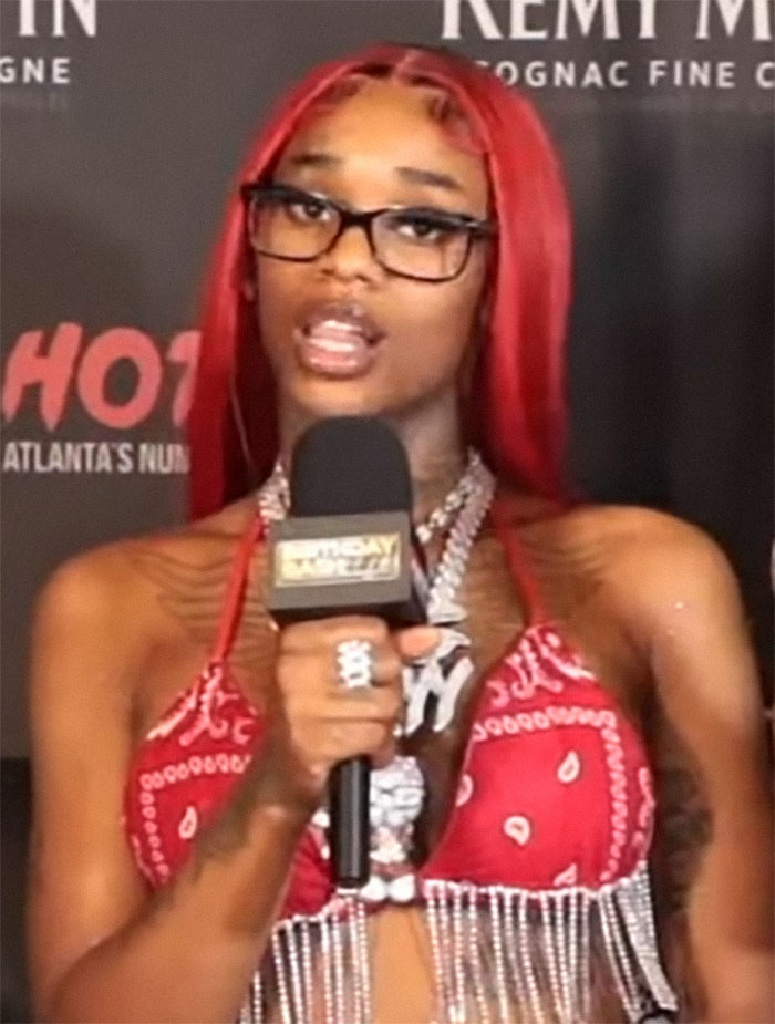 Singer in a red outfit and glasses holding a microphone at an event backdrop.