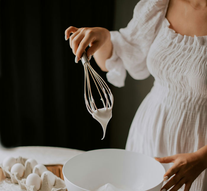 A woman in a white dress using a whisk to make meringue in a kitchen setting.