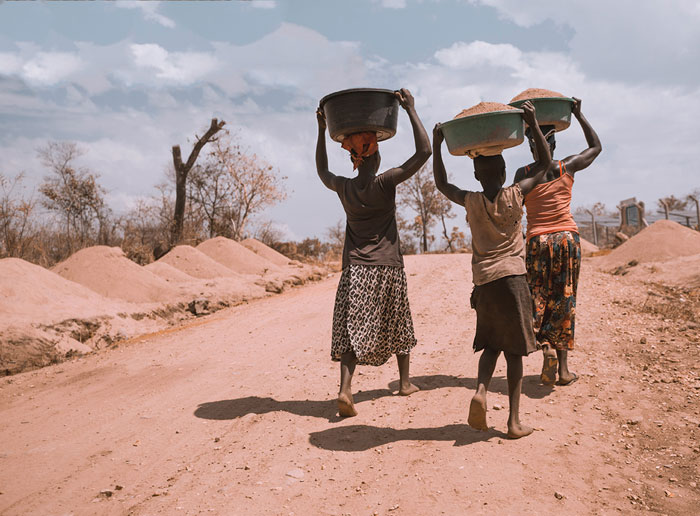 Women carrying baskets on their heads, walking on a dirt road, symbolizing contrast with rich people's out-of-touch lifestyles.