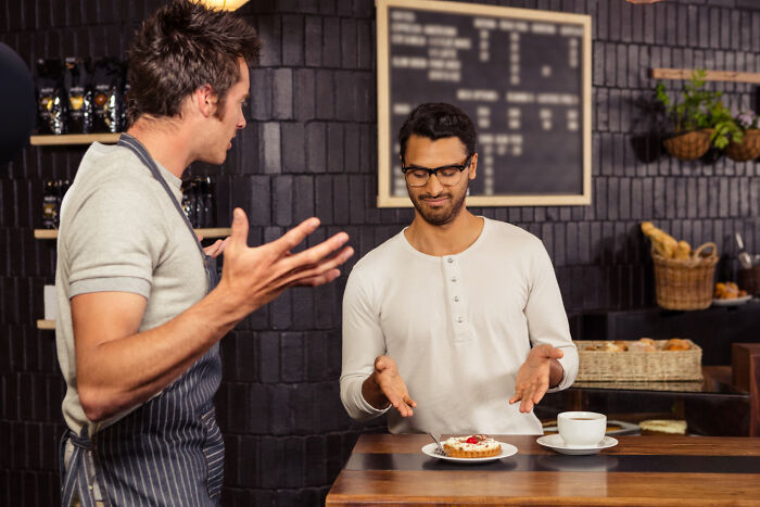 Barista and customer discussing non-physical qualities at a trendy cafe counter, with a pastry and coffee in view.