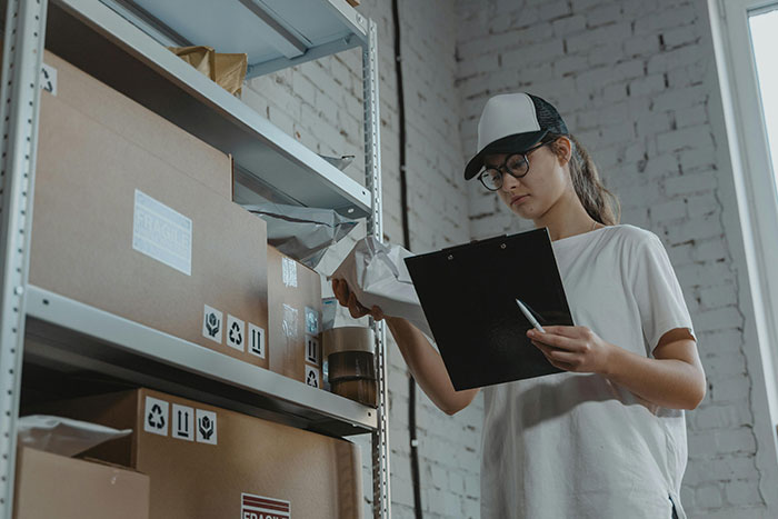 Woman organizing boxes on shelves, holding a clipboard, as she explores safe side hustle ideas.