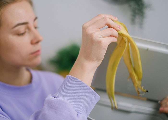 Person in a purple sweater holding a banana peel near a trash bin, focusing on waste disposal.