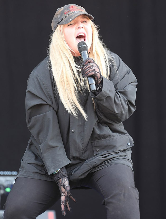 Singer performing energetically on stage, wearing a black outfit and cap, holding a microphone.