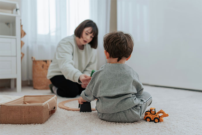 Woman playing with a child on a carpet, with toys and a basket in the background, related to babysitting.