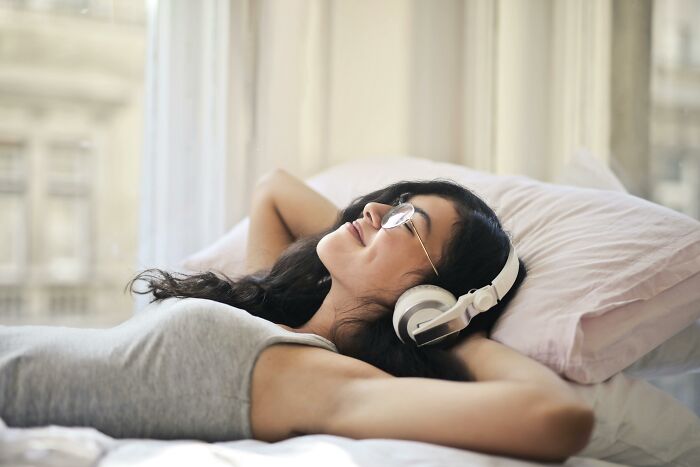 Woman with glasses and headphones relaxing on bed by window, representing things associated with rich people.
