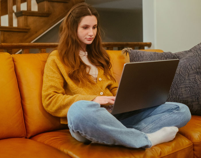 Woman in a mustard cardigan on a couch using a laptop, possibly looking into a man's background.