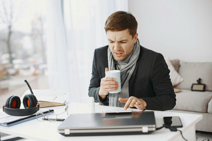 A sick man working from home, holding a mug, surrounded by work items, showing perks of not going to the office.