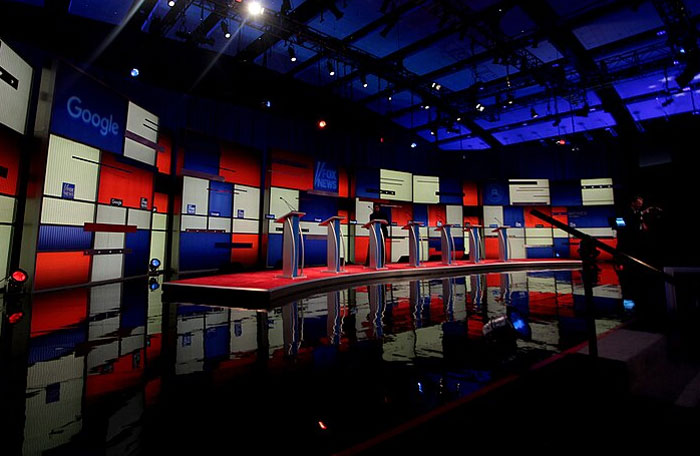 Debate stage with podiums and colorful backdrop, showcasing themes of wealth and reality.