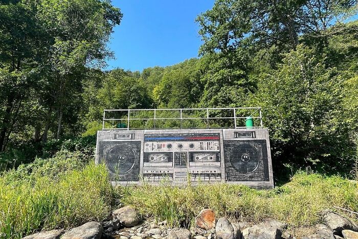 Mural by Jan Is De Man featuring a hyperrealistic boombox illusion on a city wall surrounded by lush greenery.
