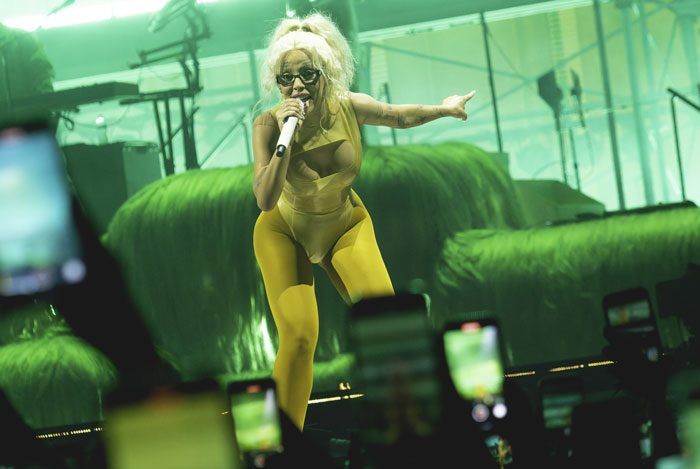 Singer performing energetically on stage in green lighting, audience recording on phones.