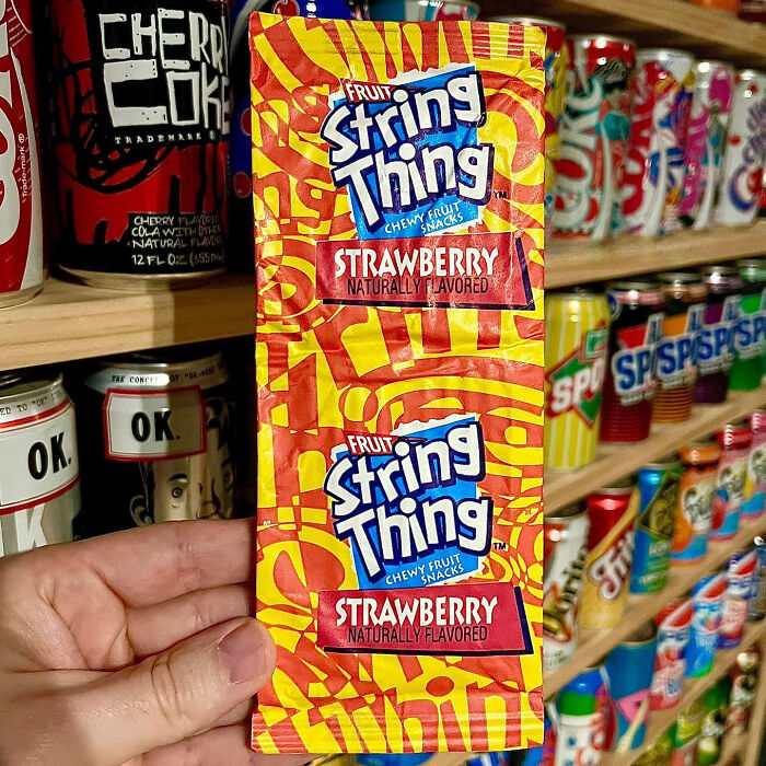 90s and 2000s food item, Fruit String Thing, strawberry flavor, held in front of retro soda cans display.