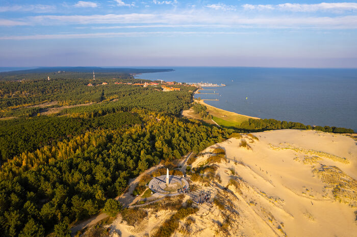 Bird’s-eye view of Lithuania showcasing sandy dunes, lush forests, and a distant coastline under a clear blue sky.