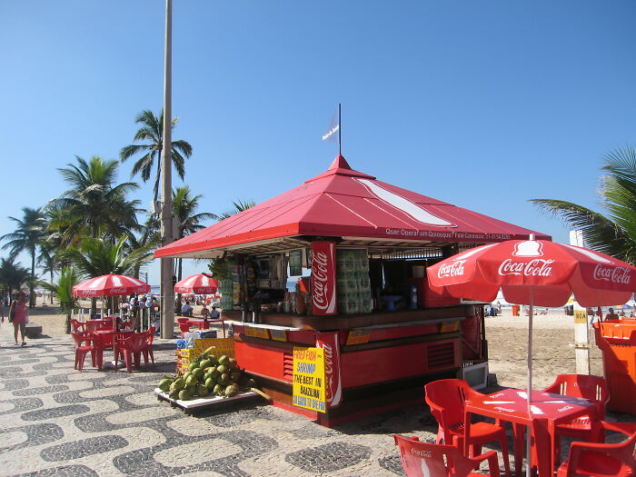 Retro Brazilian beach kiosk with Coca-Cola umbrellas, surrounded by palm trees and sunlit sand.