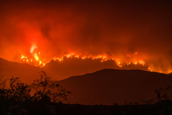 Wildfire spreading across a mountain range at night, with intense flames and smoke illuminating the scene.
