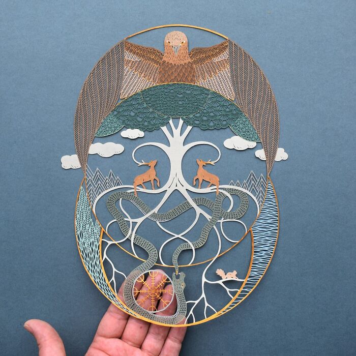 Intricate paper art design inspired by nature, featuring animals and a tree, held by a hand against a blue background.