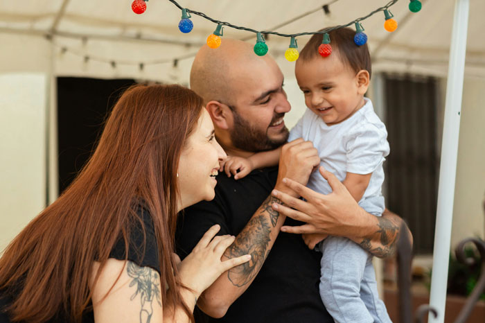 A cute family smiling and playing with their child under colorful string lights at a restaurant.